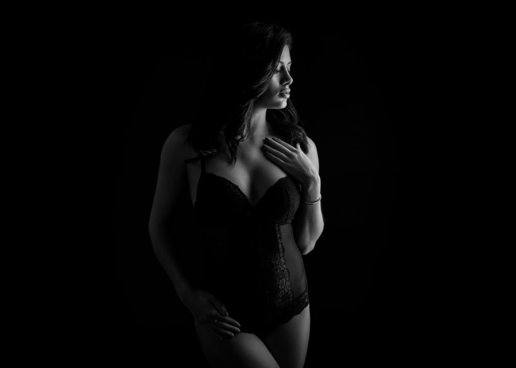A woman in black lace lingerie stands against a dark background, softly lit from the side, looking away and touching her chest with one hand. The lighting creates a dramatic, elegant effect, perfect for a beauty portrait session.