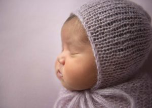 A close-up side profile of a sleeping baby wearing a soft, knitted lavender bonnet and wrap, against a light purple background.