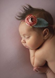 A sleeping baby lies on a soft pink surface, wearing a headband with a pink flower and green leaf. Captured by NYC's top-rated newborn photographer, the baby's hair is styled in a small, upright tuft.