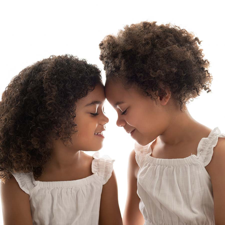 Two young sisters face each other, with their foreheads touching. Their facial profiles are silhouetted by a white backdrop in this pleasant studio portrait