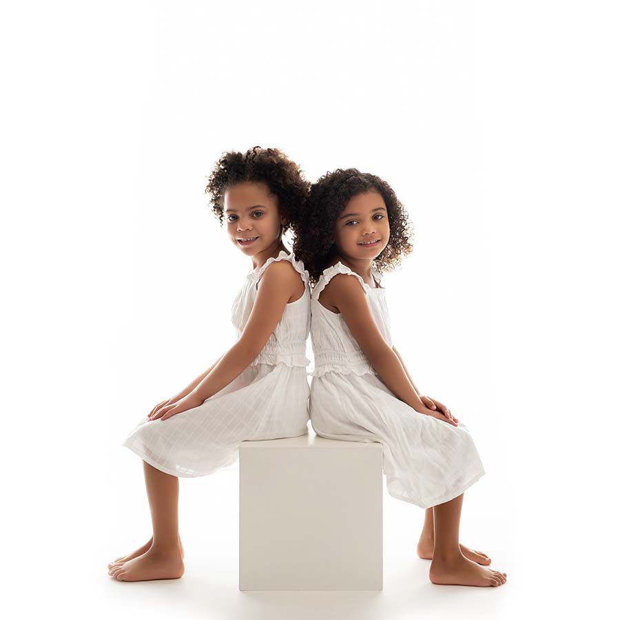 Two sisters dressed in white dresses sit on a posing block at a NYC photo studio. They are sitting back to back, with happy expressions on their faces.