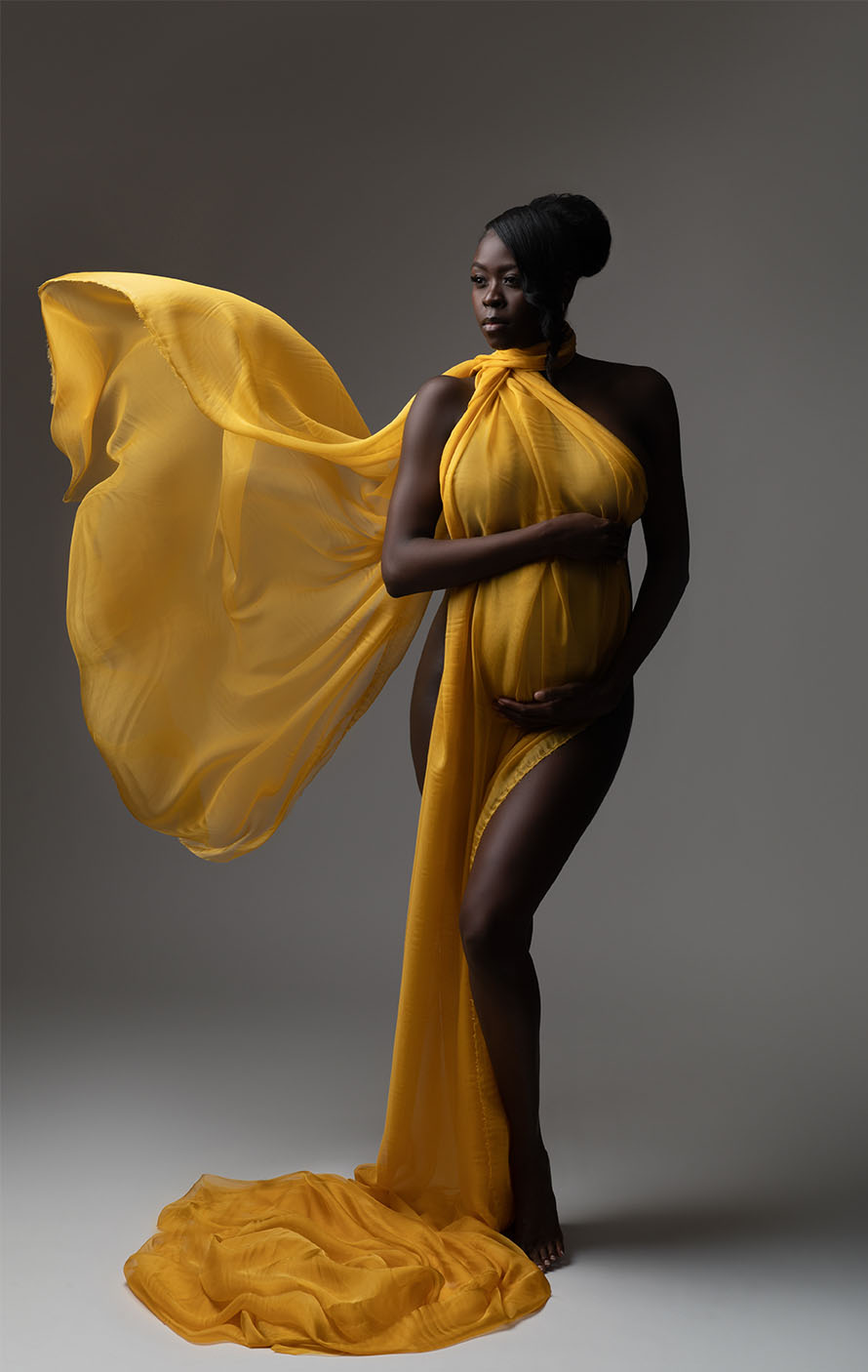 A pregnant woman stands confidently in a photo studio, wrapped in a flowing yellow fabric that billows dramatically behind her. She holds her baby bump with both hands, standing against a neutral background, with gentle lighting highlighting her serene and strong pose.