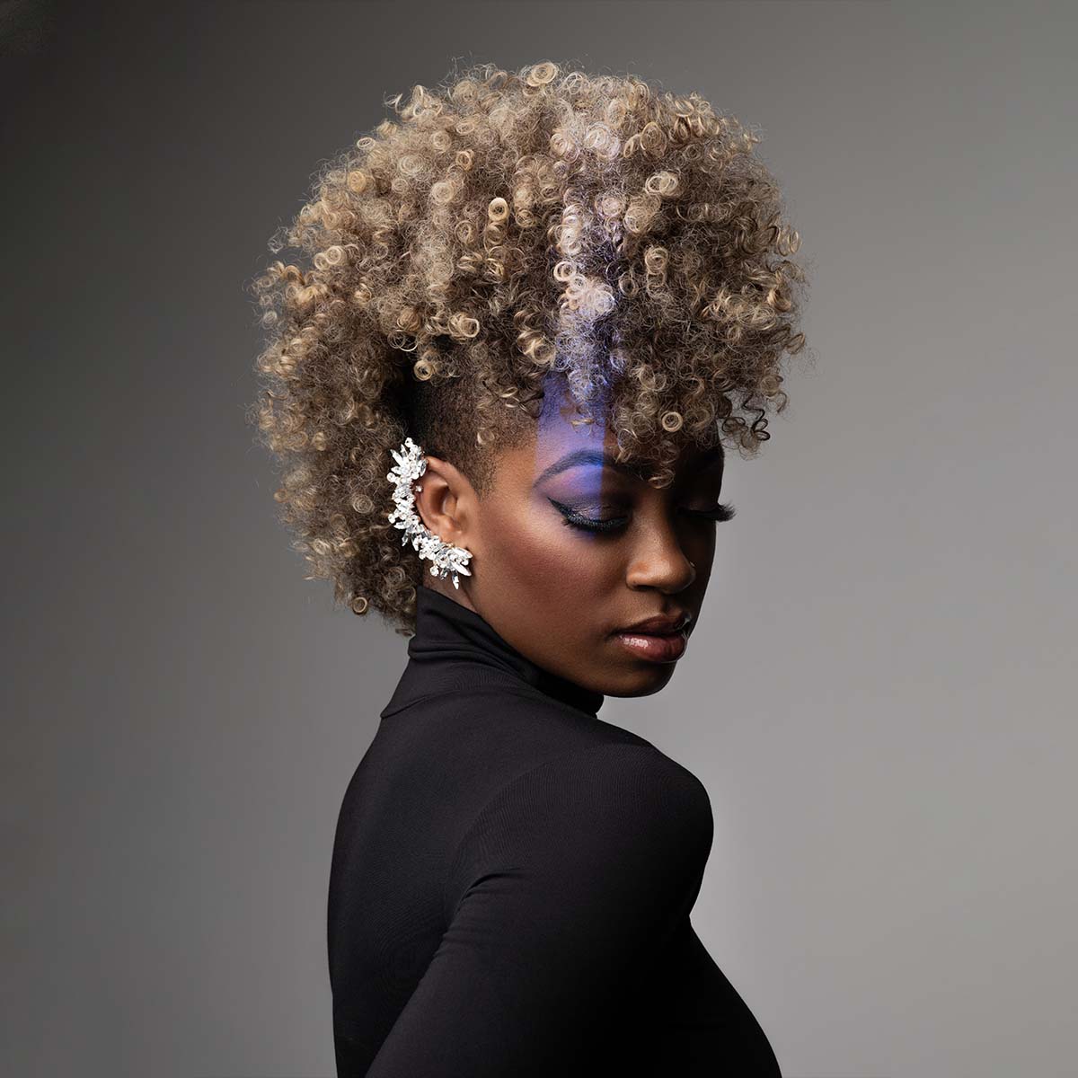 A woman with curly, blonde hair styled in a mohawk poses against a gray background. She is wearing a black turtleneck and large, decorative earrings. Her eyes are closed, and her makeup includes vibrant blue eyeshadow.