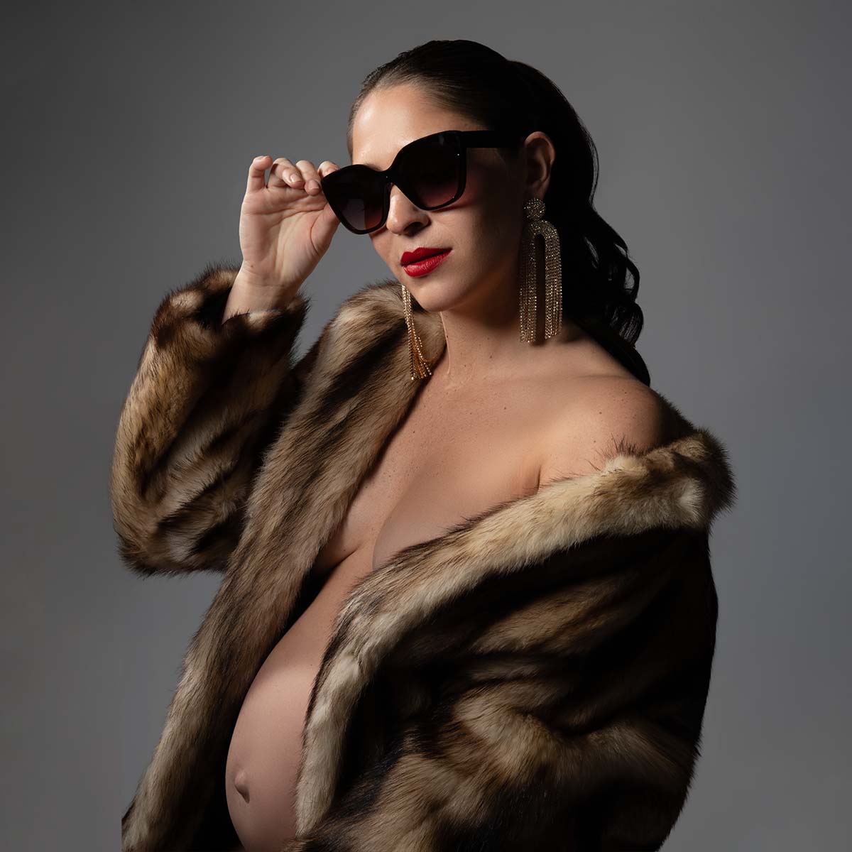 A pregnant person wearing a fur coat, sunglasses, and large dangling earrings poses confidently against a plain background. The coat is draped over their shoulders, showcasing their bare torso and visible baby bump.