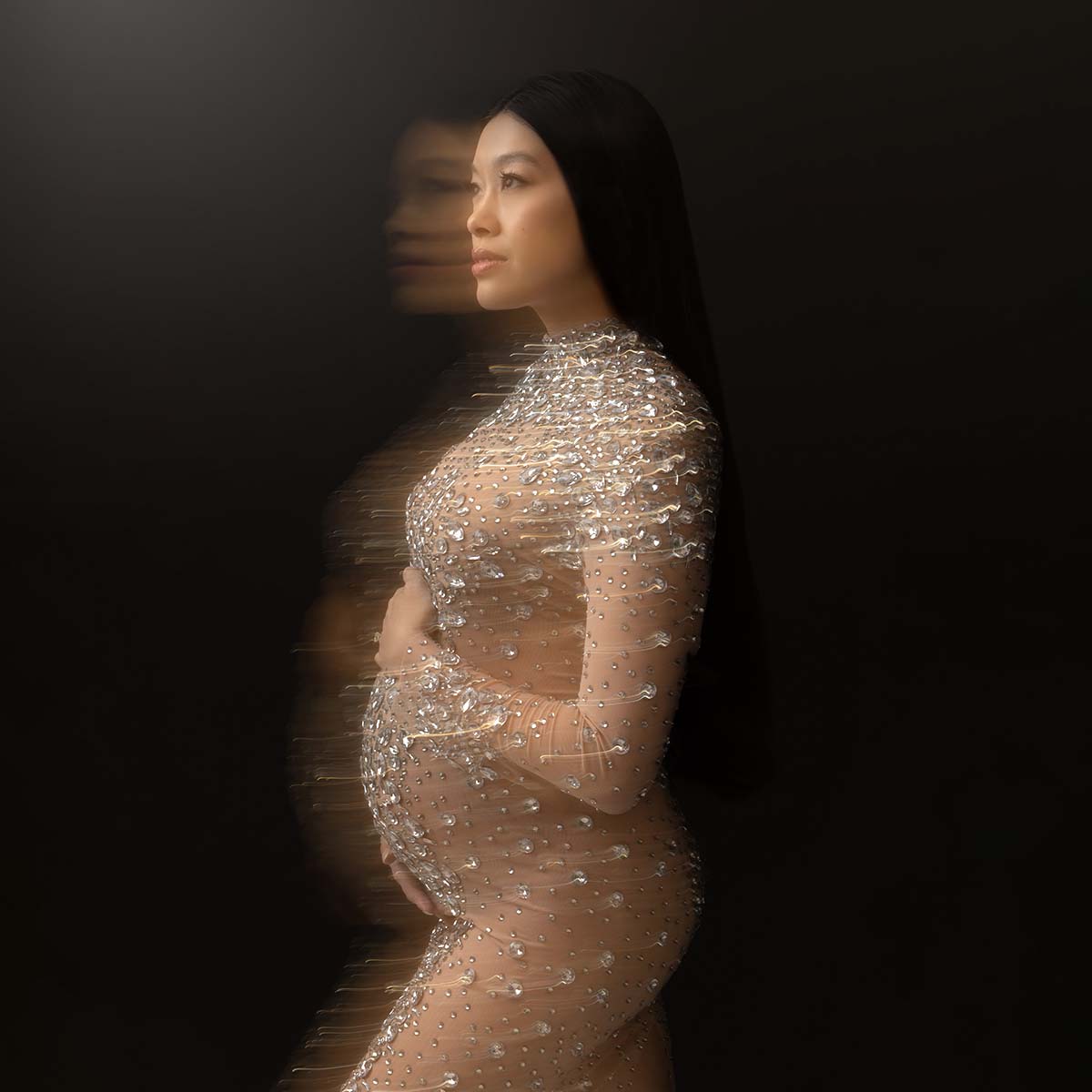 Pregnant woman in a sparkling, translucent dress poses against a dark background. Perfect for maternity photography, the dress is adorned with glittering embellishments, and a motion blur effect creates a dreamy, ethereal appearance. She has long dark hair.