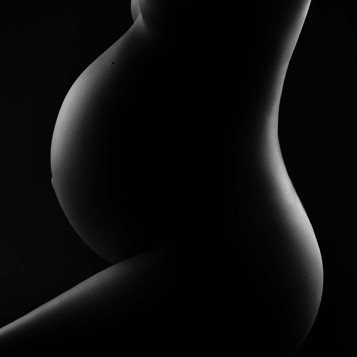 Silhouette of a pregnant person's midsection, highlighting the curve of the belly in a dark, monochromatic setting. The lighting emphasizes the contours against a black background.