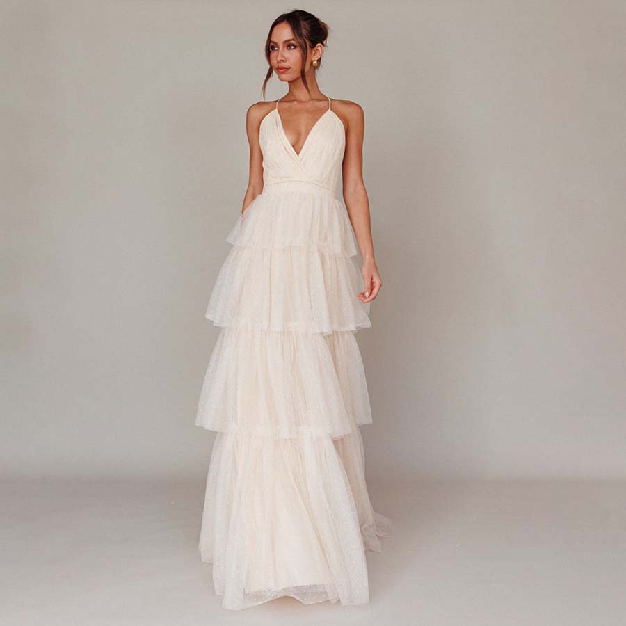 A woman stands wearing a tiered, sleeveless, cream-colored gown with a deep V-neckline. The dress features multiple layers of tulle, creating a flowing, elegant look. She is against a plain, neutral background.