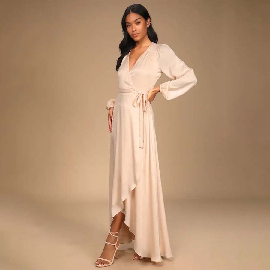 A woman stands confidently wearing a long, elegant cream dress with a deep V-neckline and long sleeves. The dress has a wrap design with a tie on the side and an asymmetrical hem. She is also wearing strappy high heels. The background is plain beige.