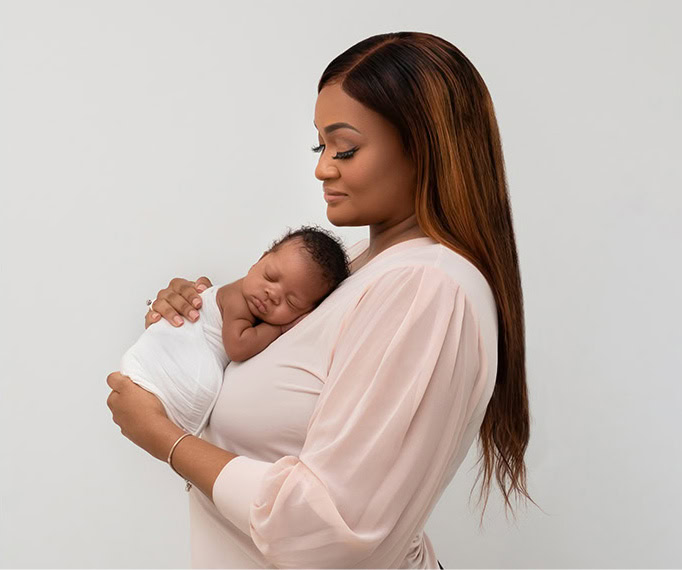 An edited version of a portrait shows a woman with long hair in a pale pink top holding a sleeping newborn, wrapped in a white blanket against her chest, both facing left against a plain light background.