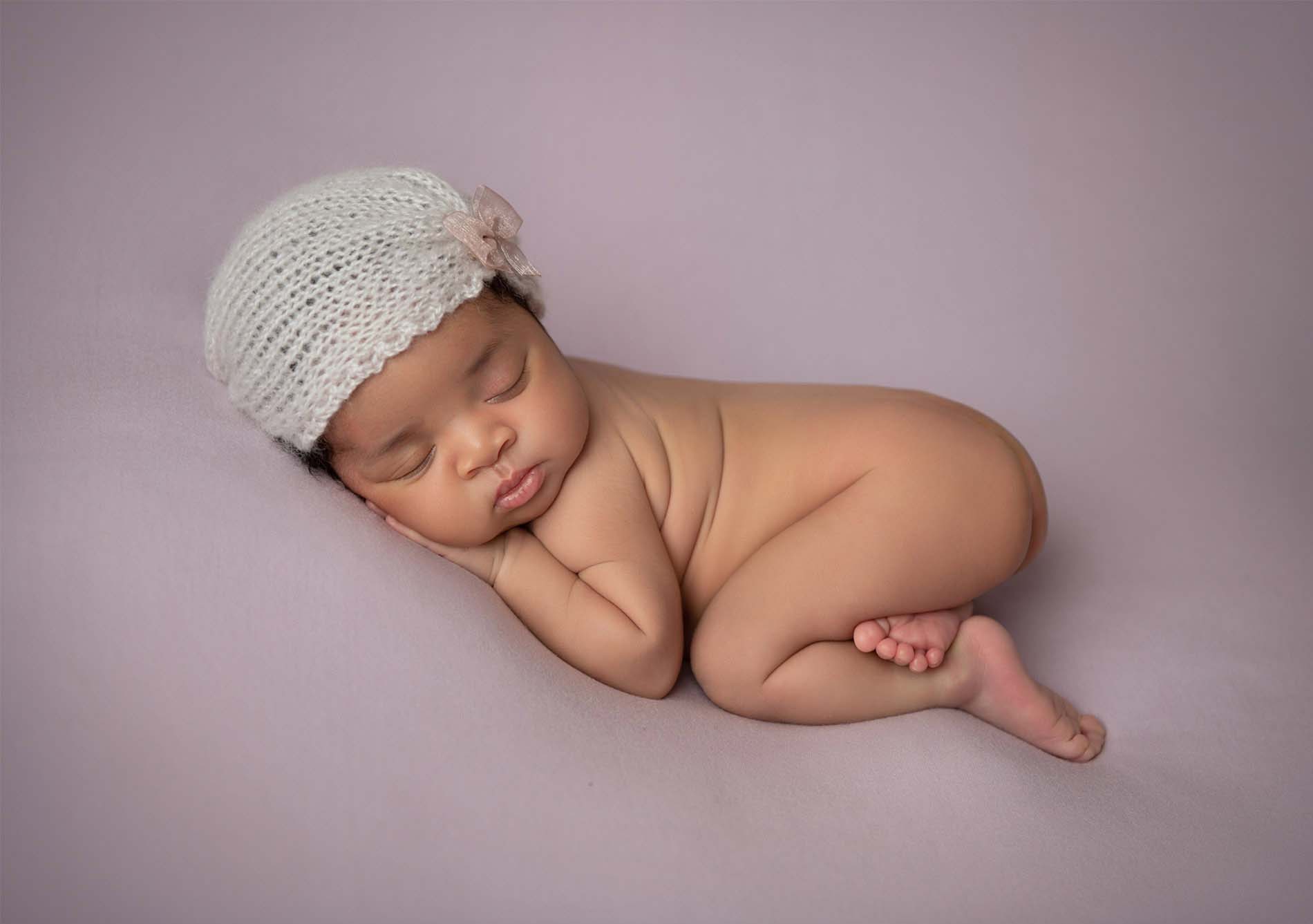 A newborn baby sleeps peacefully on a soft, light purple blanket, wearing a white knit hat with a small bow, with one hand under their cheek and legs curled up.