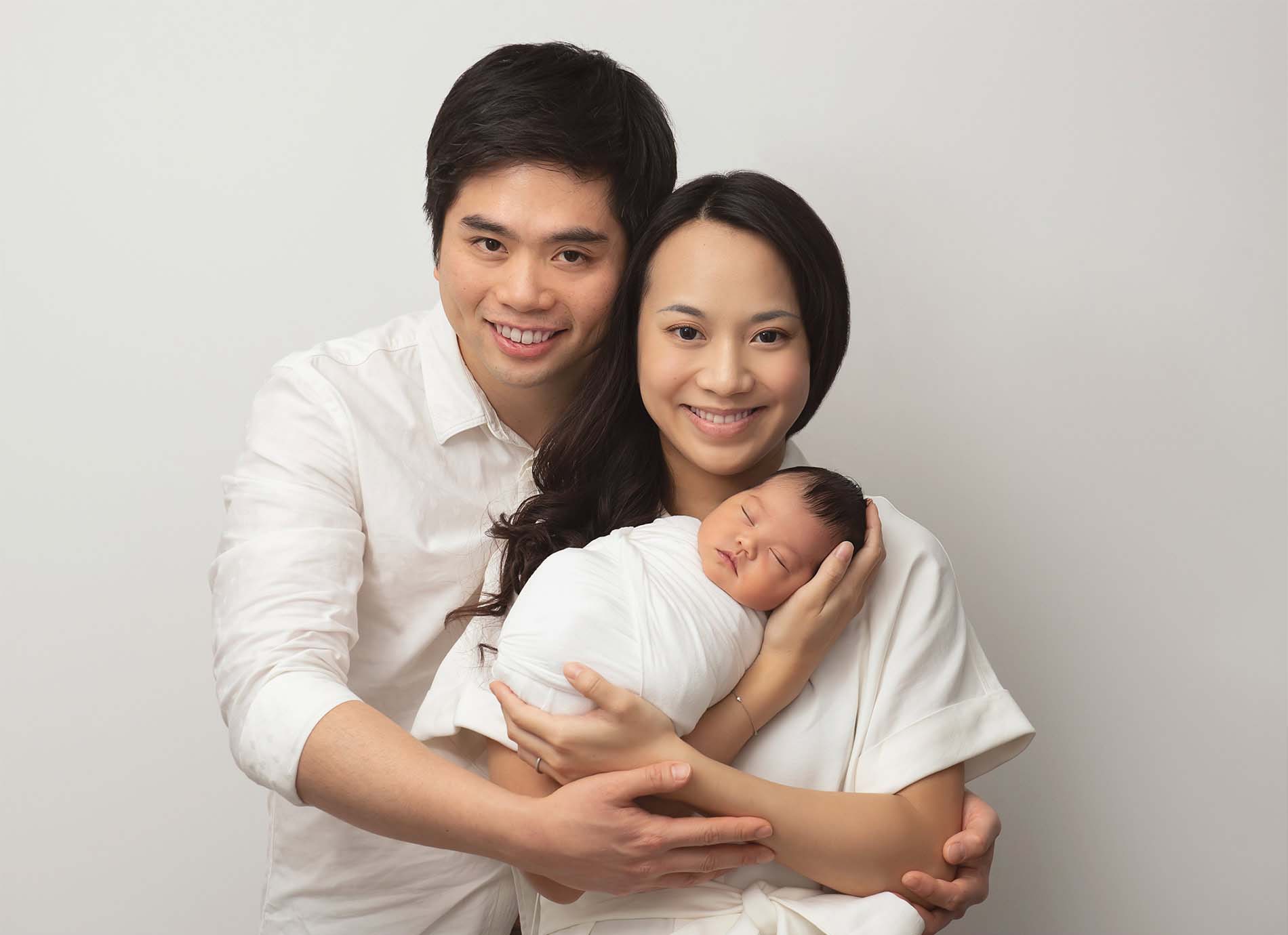 A smiling couple poses together, holding a newborn baby wrapped in a white blanket. The family stands against a plain, light-colored background, looking happy and content.
