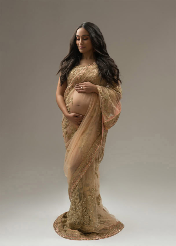 A pregnant woman in a gold embroidered sari stands against a plain background, gently cradling her baby bump and looking down with a serene expression, capturing a beautiful moment in her prenatal photoshoot.