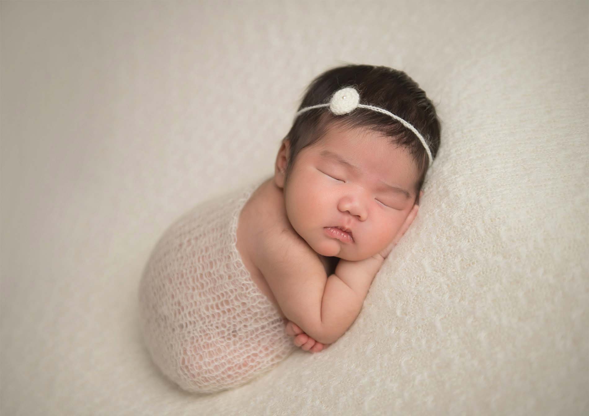 A newborn baby sleeps peacefully on a soft, light-colored blanket, wrapped in a delicate knitted wrap and wearing a simple headband with a small white flower.