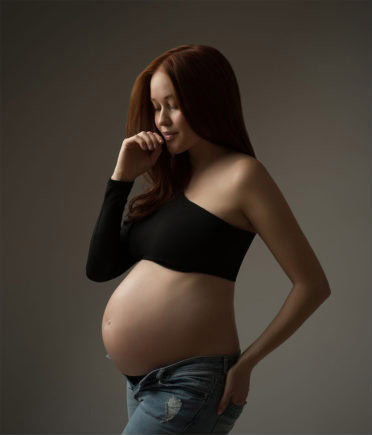 A pregnant woman with long brown hair stands in profile, wearing a black off-shoulder top and unbuttoned jeans. She gently touches her face and her belly, looking down with a soft expression against a plain background.