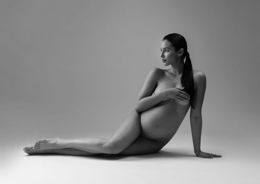 A pregnant woman poses nude in a studio, reclining on her side with one arm extended behind her for support and the other hand covering her chest. The image is in black and white with a plain background.