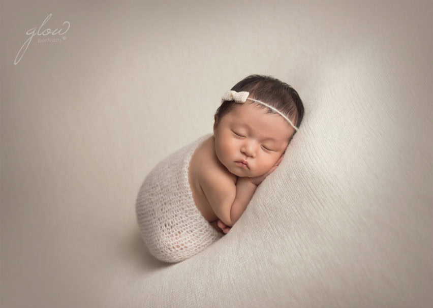 A newborn baby sleeps peacefully on a soft, cream-colored surface, wrapped in a knitted white blanket, with a small white headband on her head. The background is smooth and neutral.