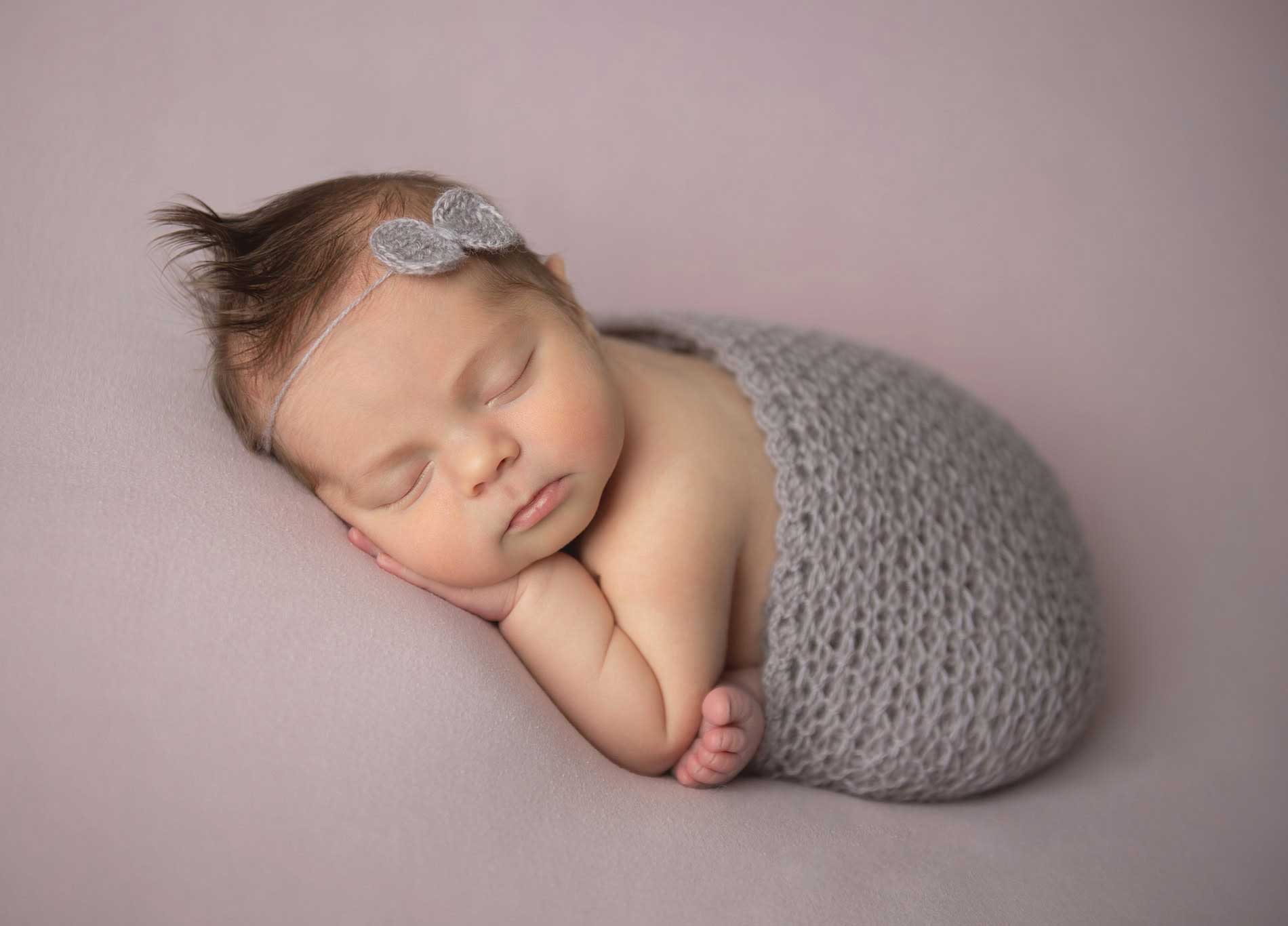 A sleeping newborn baby lies on a soft, light purple blanket, swaddled in a gray knit wrap, with a gray bow headband. The baby rests one cheek on their arm, with peaceful expression and slightly tousled hair.