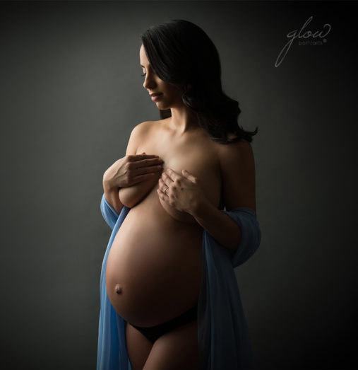 A pregnant woman stands against a dark background during a pregnancy photoshoot at a NYC studio, wearing blue sheer fabric draped over her shoulders and black underwear. She covers her chest with her hands, softly lit to highlight her baby bump.