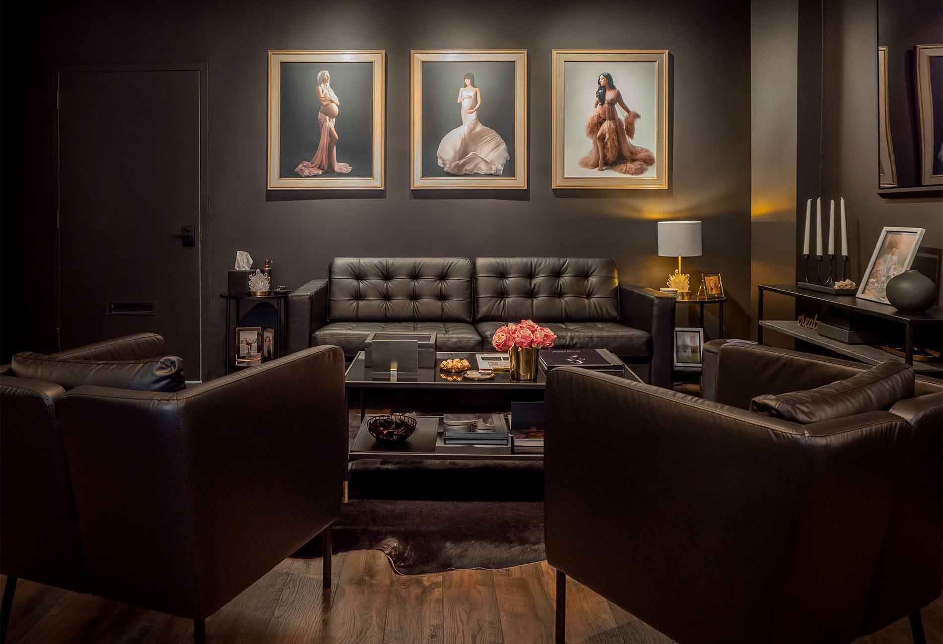 A modern, elegant living room inside of a photo studio features black leather chairs and sofa, a glass coffee table with flowers and snacks, dark walls, framed portraits of women in gowns, and soft, warm lighting.