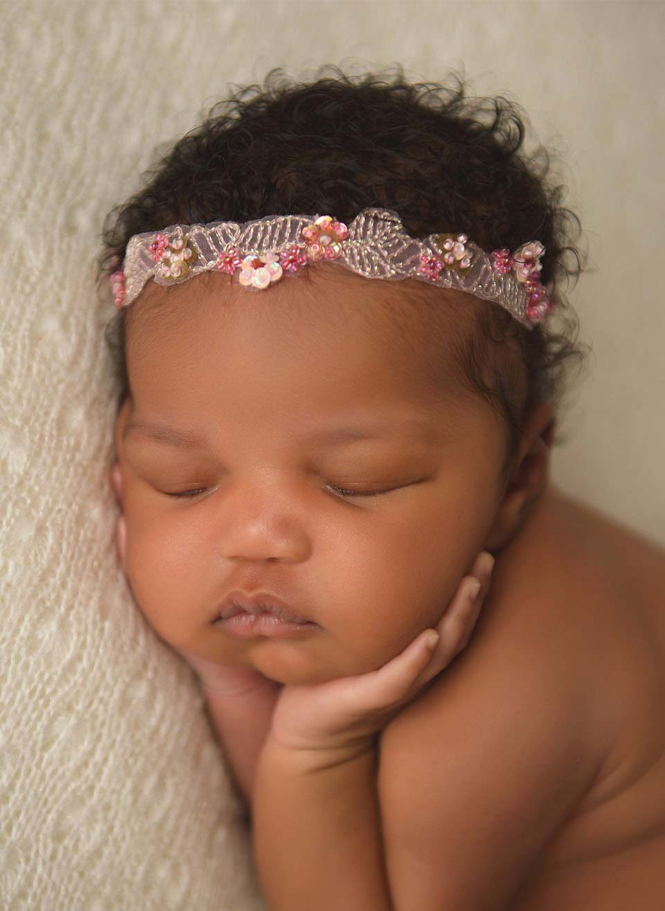 A newborn baby with curly hair and a delicate floral headband sleeps peacefully on a soft, light-colored blanket, resting their chin in their hands—capturing the gentle magic of newborn photos at home.