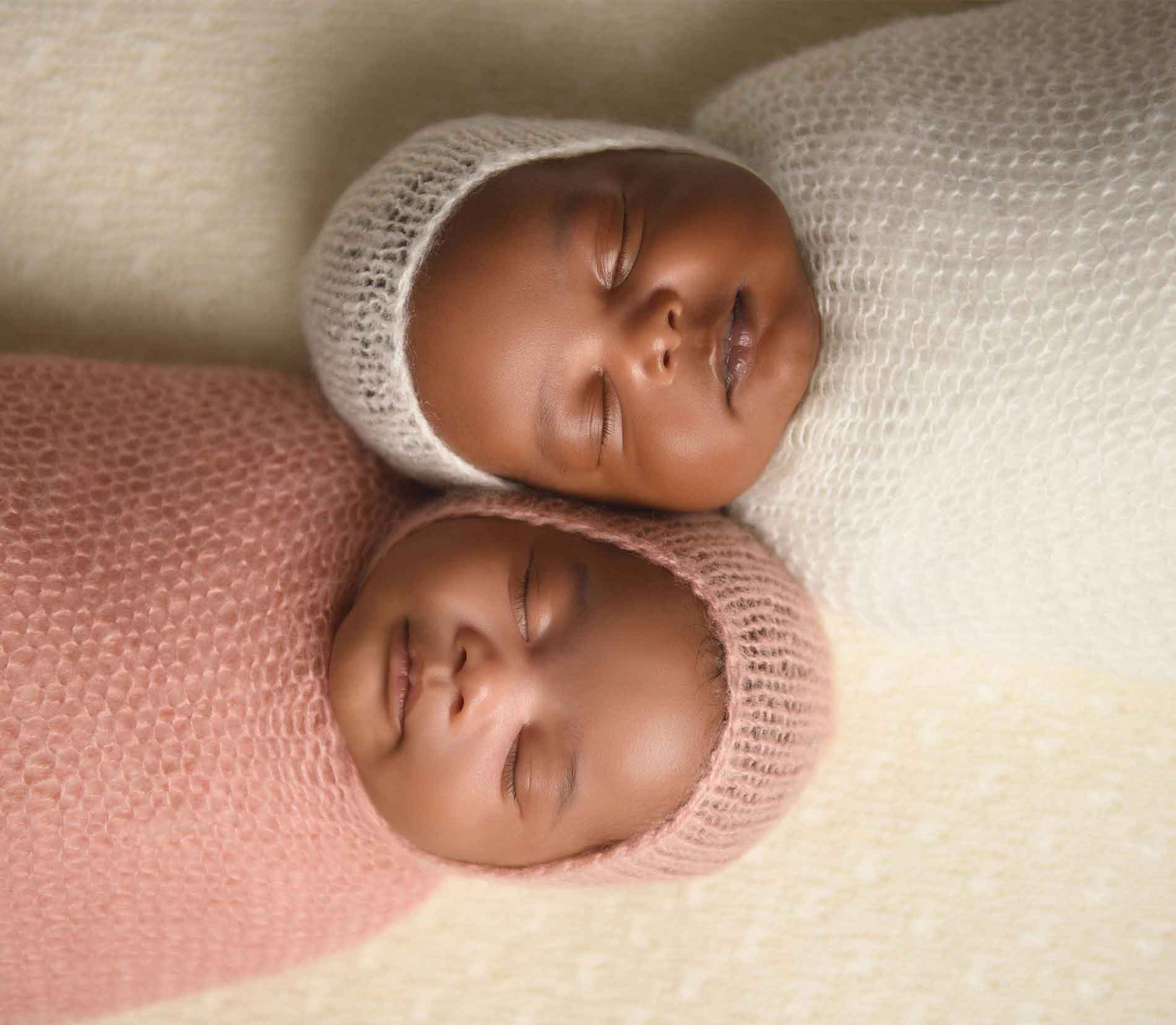 Two newborn twins sleep side by side, wrapped snugly in knitted pink and white blankets with matching hats, capturing a precious moment perfect for a newborn twins photoshoot on a soft, light-colored surface.