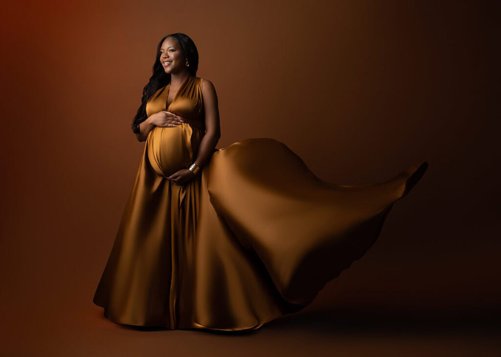 A pregnant woman in a flowing, golden-brown dress stands smiling in an NYC photo studio against a brown background, holding her belly with one hand and her chest with the other as the dress billows dramatically to one side.