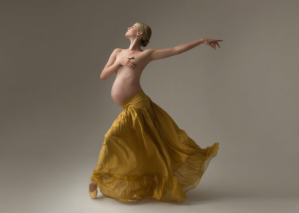 A pregnant woman in a long, flowing yellow skirt poses gracefully on pointe during a maternity photoshoot, with one hand on her chest and the other arm extended, set against a neutral background.