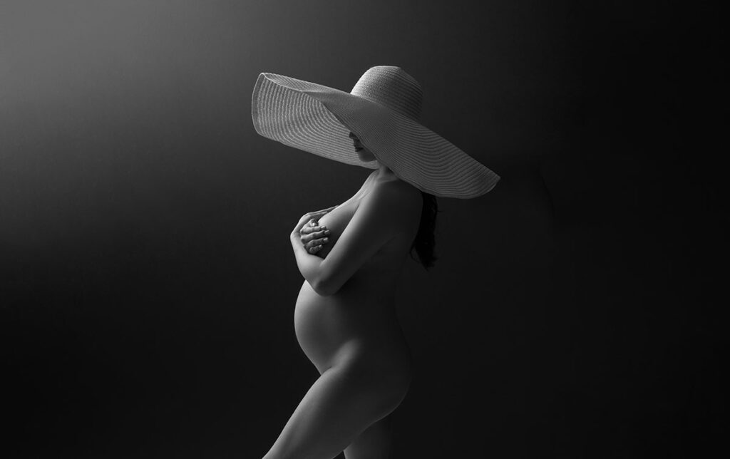 Black and white photo by a professional maternity photographer shows a nude pregnant woman in profile against a dark background, wearing a large wide-brimmed hat and covering her chest with her arms.