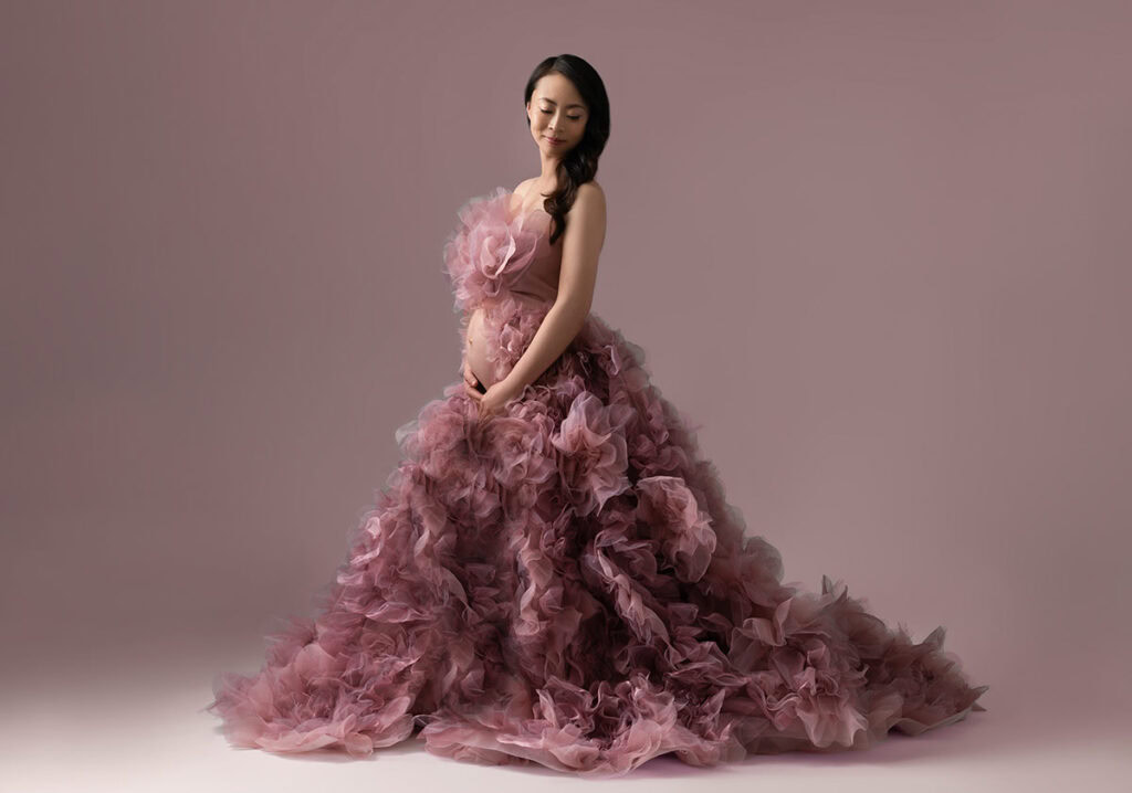 A pregnant woman in a flowing, ruffled mauve gown stands against a soft, pinkish background, gently cradling her belly and looking down with a serene expression.