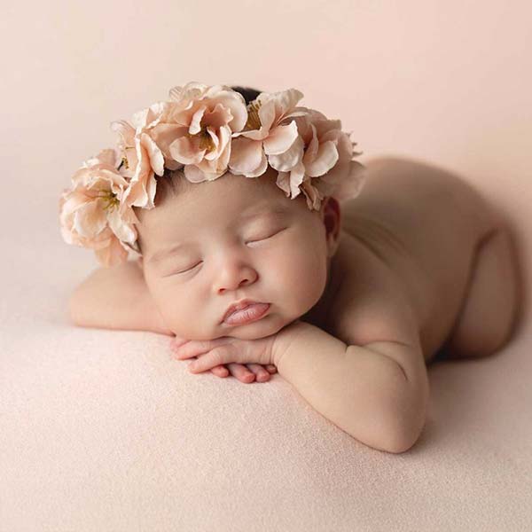 A sleeping baby rests on a soft pink surface, arms folded under their head and wearing a delicate pink flower headband. Captured in an NYC photography studio, the gentle scene is wrapped in soothing shades of pink for a timeless baby portrait.