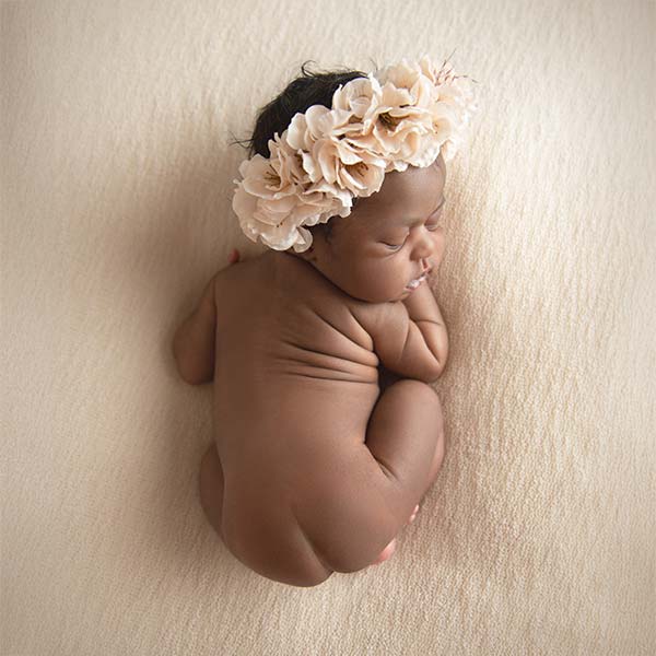 A newborn baby lies on a soft beige blanket, curled up on their side with eyes closed, wearing a large cream-colored flower headband—perfect for lifestyle infant photos in your home in NYC.