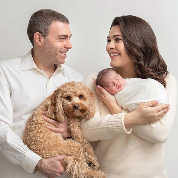 A stylish Manhattan NYC family is captured as a man, holding a small curly-haired dog, stands beside a woman cradling their swaddled newborn. Both smile warmly at each other against a neutral background.